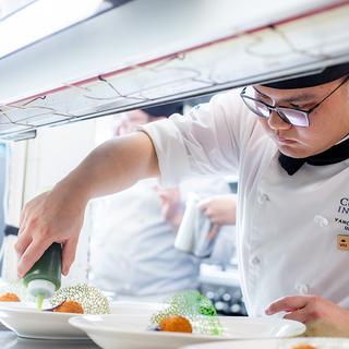 Man in chef's whites pouring sauce on a plate of food. 