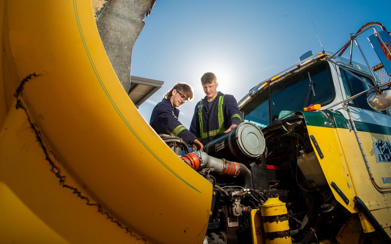 Two men working on the engine of a big yellow truck under a sunny blue sky. 